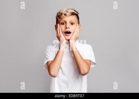 Überraschter Junge mit Händen auf dem Gesicht, der in die Kamera blickt, emotional schockiertes Kind, das Staunen und Erstaunen auf grauem Hintergrund ausdrückt, Studio-Porträt Stockfoto