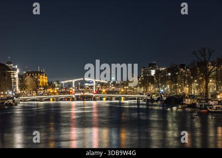 Nächtlicher Panoramablick auf die Amstel und den Kanal in Amsterdam, mit historischen Gebäuden und Brücken beleuchtet, die sich auf der dunklen Wasseroberfläche spiegeln Stockfoto
