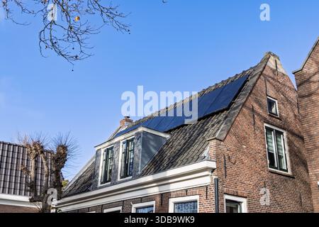 Solarpaneele, die an einem sonnigen Tag auf dem dunklen Ziegeldach eines traditionellen, monumentalen niederländischen Backsteingebäudes vor einem hellblauen Himmel installiert wurden. Stockfoto