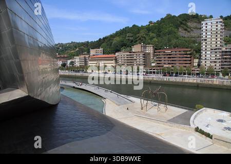 Der Fluss Nervion (Ría de Bilbao) fließt vorbei am berühmten Guggenheim Museum in Bilbao, Biskaya, Spanien. Die avantgardistischen Titankurven von Frank Gehrys architektonischem Meisterwerk befinden sich am linken Ufer und spiegeln sich im Wasser. Diese malerische Aussicht fängt das Herz des Baskenlands in den industriellen und kulturellen Wandel und die moderne Uferpromenade der Stadt ein. Stockfoto