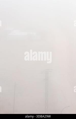 Ein vertikales Bild, bei dem die Oberseite eines Power-Pylons die untere Hälfte dominiert, während schwache, rechteckige Strukturen, möglicherweise Gebäude, kurz daraus hervorgehen Stockfoto