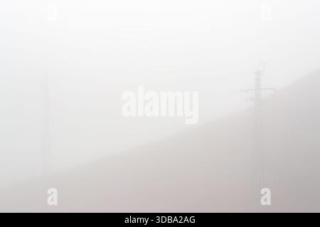 Ein schlichtes, minimalistisches Bild eines nebelbedeckten Hügels mit einer sichtbaren Stromleitung, die zwischen zwei weit entfernten Sendetürmen verläuft Stockfoto