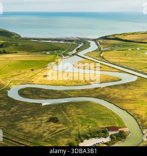 Aus der Vogelperspektive auf einen Serpentinfluss, der sich durch pulsierende grüne Felder schlängelt, in Richtung des schimmernden Meeres, ein kleines Gebäude in der Nähe seines Ufers, Eastbourne, Stockfoto