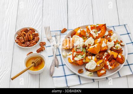 Gebackene Butternusskürbscheiben mit Mozzarella, zerbröckeltem Käse, Thymian, gerösteten Pekannüssen auf Teller auf weißem Holztisch mit Gabeln, Honig, horizontale Ansicht Stockfoto