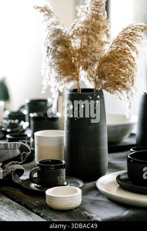 Rustikale Tischgestaltung mit leerem handgemachtem Keramikgeschirr, schwarz-weißen groben Schüsseln, Tassen, Kanne und Vase auf Leinentischdecken. Trockene Schilfblume Stockfoto