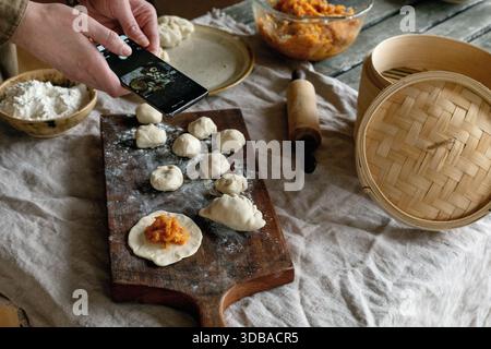 Asiatische gedämpfte Teigtaschen mit Gemüsefüllung herstellen. Weibliche Hände Formen Knödel auf bemehltem Brett, Dampfgarer und Zutaten in der Nähe. Natürliches Tageslicht Stockfoto