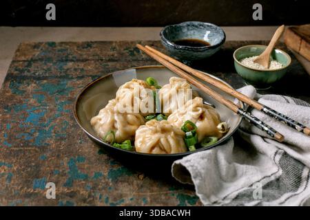 Hausgemachte asiatische gedämpfte Teigtaschen in grauer Platte, belegt mit Sesamsamen und grünen Zwiebeln. Sojasauce, Essstäbchen, graue Serviette in der Nähe auf rustikalem Holz B Stockfoto
