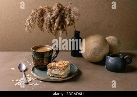 Hausgemachter norwegischer süßer Kuchen Verdens Beste Kake mit Vanillepudding, Meringue und Mandeln, ein quadratisches Stück auf Teller auf beigem Tisch mit Kaffeetasse, Löffel, Stockfoto
