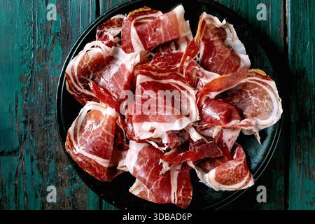 In dünne Scheiben geschnittener spanischer Schinken Jamon Iberico von der Schweinescheibe paleta auf schwarzem Teller. Alter hölzerner Hintergrund, dunkles Türkis, Blick von oben. Kopierbereich Stockfoto