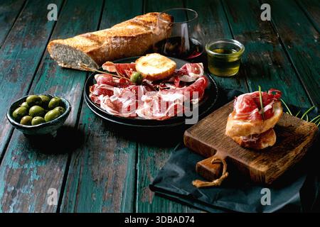 In Scheiben geschnittener spanischer Schinken Jamon Iberico paleta auf schwarzem Teller. Schüssel grüne Oliven, frisches und geröstetes Weißbrot, Olivenöl, Glas Rotwein, Tapas. Alt Stockfoto