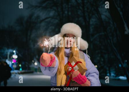 Junge Frau mit Pelzmütze und warmer Kleidung, mit einem glitzernden Funkeln und einem umhüllten Geschenk, die bei den nächtlichen Winterfeiern eine festliche Atmosphäre schafft Stockfoto