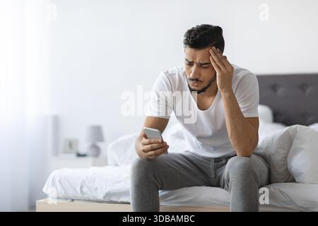Verärgerter arabischer Mann liest schlechte Nachrichten auf dem Smartphone, während er auf dem Bett im Schlafzimmer sitzt Stockfoto