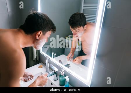 Junger Mann, der Schaum am Spiegel im kompakten Badezimmer spült, sich über Waschbecken mit fokussiertem Ausdruck beugt, lässiges Handtuch in der Nähe, Pflege || Model veröffentlicht Stockfoto