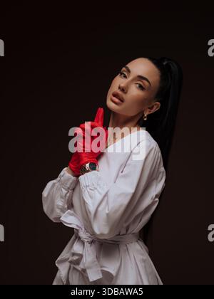 Selbstbewusste junge Ärztin oder Krankenschwester Kosmetologin weiß Uniform rote Handschuhe Zeigefinger. Professionelle ästhetische Aufmerksamkeit und Expertise Stockfoto