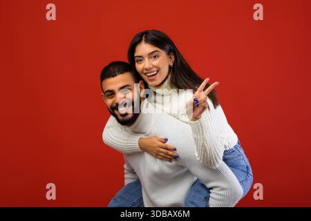 Fröhliches Arabisches Paar Genießt Die Zeit Zusammen, Guy Gibt Der Frau Huckepack-Fahrt Mit Friedenszeichen Geste Stockfoto