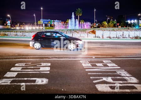 Eine Low-Winkels-Aufnahme auf Straßenniveau, bei der ein schwarzes Auto nachts über eine Stadtstraße fährt und einen Bewegungsunschärfeeffekt erzeugt. Im Hintergrund ein helles Licht Stockfoto