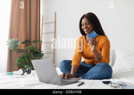 Junge Frau beim Online-Shopping mit Kreditkarte, während sie in einem gemütlichen Zimmer auf ihrem Bett sitzt Stockfoto