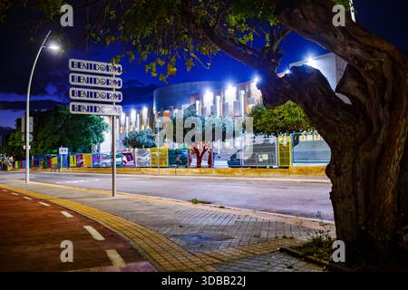 Eine breitere, aus einem tiefen Winkel gefertigte Aufnahme bei Nacht, die den Richtungsschild „BENIDORM“ zwischen einem großen, strukturierten Baumstamm im Vordergrund und einem b zeigt Stockfoto