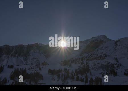Ein atemberaubender Blick auf einen Winteruntergang über schneebedeckten Bergen. Die Sonne bricht durch die Gipfel und strahlt ein warmes Leuchten über die Landschaft. Die sc Stockfoto