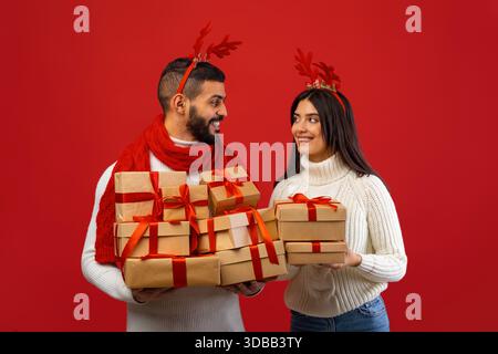 Liebevolles arabisches Paar feiert Neujahr mit Geschenken und freudvollem Lächeln auf rotem Hintergrund Stockfoto