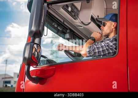 Ein Mann sitzt auf dem Fahrersitz eines roten Lkws, während er nach draußen blickt. Er trägt eine Sonnenbrille und eine Mütze. Stockfoto