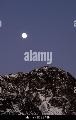 Ein atemberaubendes Foto fängt einen Vollmond ein, der über einem schneebedeckten Berggipfel hängt. Das dunkle, felsige Gelände des Berges bildet einen wunderschönen Kontrast zu Stockfoto