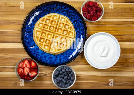 Blick von oben auf eine goldene Waffel auf einem blauen Teller, umgeben von frischen Himbeeren, Heidelbeeren, Erdbeeren und Schlagsäpfel für Brunch in Waukes Stockfoto
