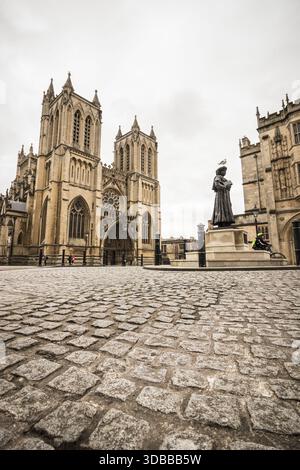 Der Blick auf die große Fassade der Kathedrale von Bristol erhebt sich majestätisch über dem kopfsteingepflasterten Platz, im Schatten einer Statue unter einem bewölkten Himmel, Bristol, eng Stockfoto