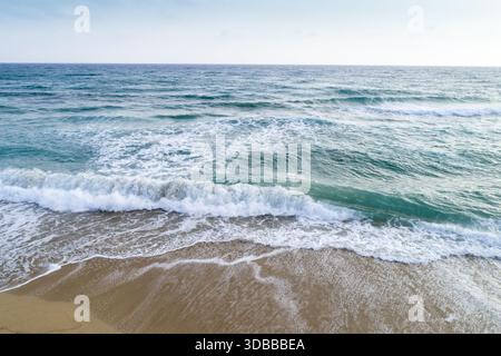 Aus der Vogelperspektive auf das türkisfarbene Meer, das den goldenen Sand küsst, Wellen, die in schaumiger Pracht unter einem ruhigen Himmel krachen, Vrachos, Preveza, Griechenland. Stockfoto