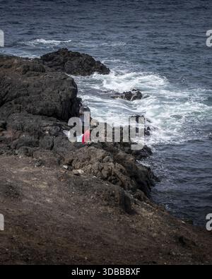 Blick auf zerklüftete vulkanische Felsen, die auf das turbulente, dunkelblaue Meer treffen, während ein Fischer seine Linie unter der hellen Sonne wirft, Fuerteventura, die Kanarischen Inseln, Stockfoto