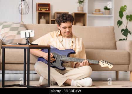 Junger glücklicher Mann mit Handy spielt neue blaue Gitarre auf dem Boden zu Hause Stockfoto