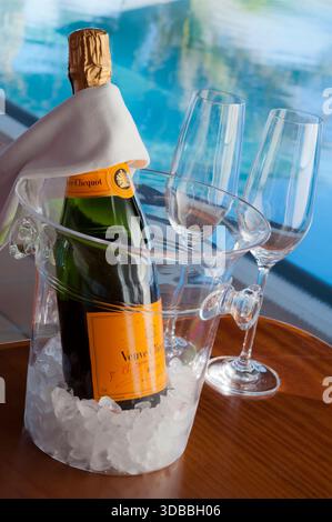 Veuve Clicquot edle Vintage-Champagnerflasche mit Gläsern, auf Eis im Weinkühler, luxuriöser, sonnendurchfluteter Infinity-Pool dahinter mit reflektierenden Palmen Stockfoto