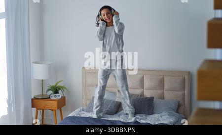 Kaukasische Frau hört Musik in den Kopfhörern auf dem Bett springen orthopädische Matratze haben Spaß um weibliches Mädchen im Schlafzimmer springen Freude lustig Stockfoto