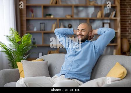 Junger Mann mit blauem Hemd und leichter Hose, bequem auf einer grauen Couch mit geschlossenen Augen und einem ruhigen Lächeln, genießt einen Moment der Entspannung und Ruhe in seinem modernen Wohnzimmer Stockfoto