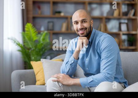 Ein kahler, bärtiger Mann sitzt selbstbewusst auf einem modernen Sofa in seinem gemütlichen Wohnzimmer, lächelt in schick-lässiger Kleidung zur Kamera, entspannt und zufrieden in einem stilvollen Innenraum Stockfoto