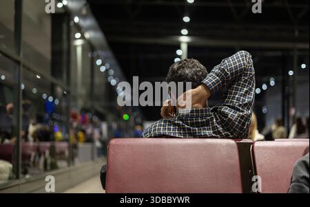 Ein einsamer Mann in kariertem Hemd wartet auf einen verspäteten Flug in einem dunklen, modernen Flughafenterminal und spricht mit der Hand hinter dem Hals am Telefon. Stockfoto