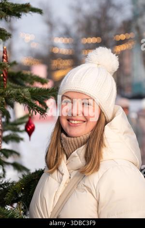 Eine lächelnde junge Frau in einer weißen Winterjacke und Strickmütze steht draußen neben einem Weihnachtsbaum mit warmen Stadtlichtern im Hintergrund Stockfoto
