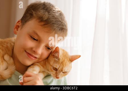 Junge lächelt und kuschelt Ingwerkatze auf seiner Schulter bei Fenster Stockfoto