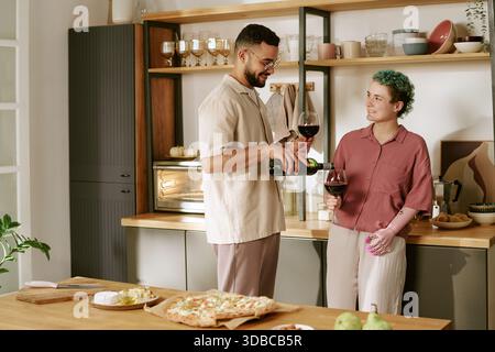 Junger Mann gießt Wein für junge Frau in der Küche lächelnd Stockfoto