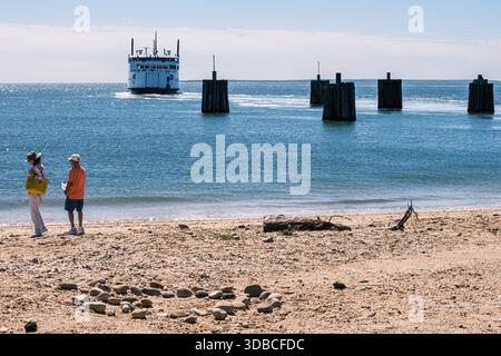Die Passagierfähre fährt von einem hölzernen Dock entlang der Küste von Long Island, während zwei Personen am Strand im Vordergrund stehen und das Wasser beobachten. Stockfoto