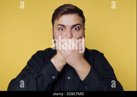 Ein Mann steht vor einem gelben Hintergrund. Seine Hände decken seinen Mund. Seine Augen sind weit offen und zeigen Überraschung oder Schock über das, was er ist Stockfoto