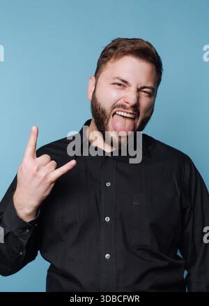 Ein Mann steht vor blauem Hintergrund. Er macht eine für Rockmusik typische Handgeste, während er seine Zunge rausstreckt. Sein Ausdruck zeigt Enthusiasmus Stockfoto