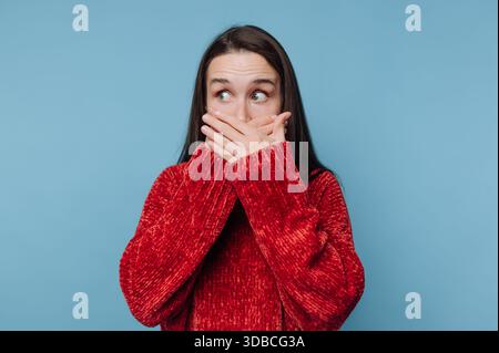Eine Frau steht vor einem blauen Hintergrund. Sie bedeckt ihren Mund mit den Händen und zeigt einen Ausdruck der Überraschung. Sie hat lange braune Haare und ist getragen Stockfoto
