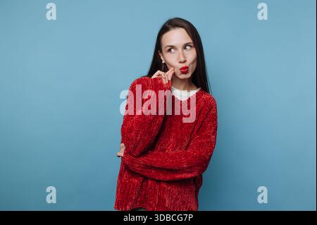 Eine junge Frau steht mit überkreuzten Armen und macht einen spielerischen Ausdruck. Sie hat lange Haare und trägt einen roten Pullover. Der Hintergrund ist ein solides blau Stockfoto