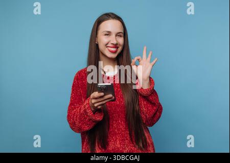 Eine junge Frau steht mit einem Smartphone in der Hand. Sie lächelt und macht mit ihrer anderen Hand eine „OK“-Geste. Der Hintergrund ist einfach blau. Stockfoto