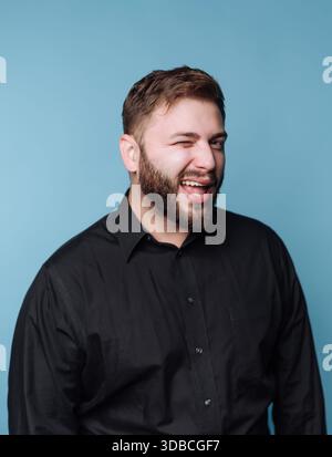 Ein Mann steht vor einem blauen Hintergrund. Er trägt ein schwarzes Hemd und lächelt, während er der Kamera zuzwinkert. Sein Ausdruck zeigt Freundlichkeit und Freude. Stockfoto