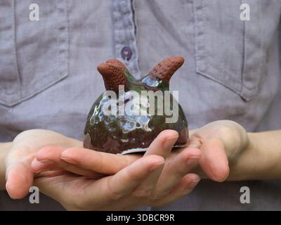 Kleiner handgefertigter Keramiktopf mit glänzend grüner Glasur und kurzen, in menschlichen Händen gehaltenen Beinen aus Ton. Nahaufnahme mit strukturierter Oberfläche und Handc Stockfoto