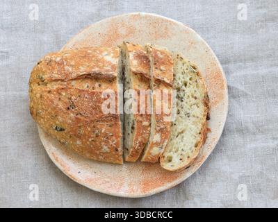 Draufsicht eines in Scheiben geschnittenen, mehrkörnigen Sauerteigbrotlaibs auf einer Keramikplatte. Verkrustete Außenseite und luftige Krümelstruktur in natürlichem Licht auf neutraler Linie Stockfoto