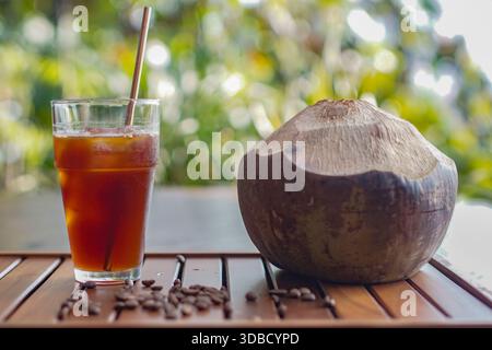 Eiskaffee serviert neben frischer Kokosnuss auf Holztisch, natürliches tropisches Getränkekonzept mit grünem Hintergrund Stockfoto