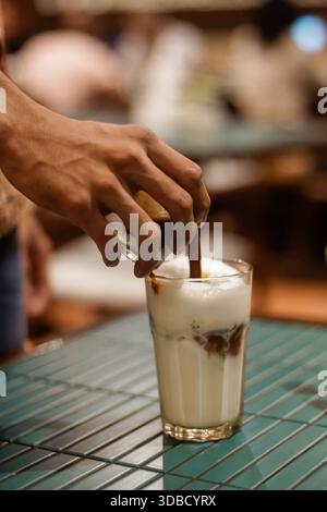 Der Barista gießt Espresso über Milchschaum in ein Eis-Latte-Glas auf der Kaffeekanne und macht den Prozess der Kaffeezubereitung aus der Nähe Stockfoto